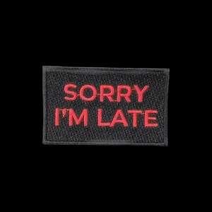 May include: A rectangular embroidered patch with a black background and a red stitched border. The text SORRY I'M LATE is embroidered in red capital letters across the centre of the patch, featuring a textured thread finish throughout.