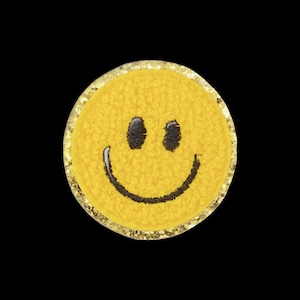 Könnte beinhalten: Ein kreisförmiger Aufnäher mit einer leuchtend gelben, strukturierten Oberfläche und einem schwarz gestickten Smiley-Gesicht. Das Design zeigt zwei schwarze, ovale Augen und einen geschwungenen schwarzen Mund, umrandet von Glitzer.