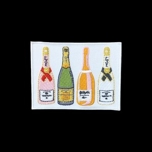 Può includere: Una toppa rettangolare ricamata su sfondo bianco raffigurante quattro bottiglie di champagne. Le bottiglie sono colorate di rosa, verde, arancione e bianco, ognuna con etichette dettagliate e nastri sul collo. Il testo MOET è visibile sulle bottiglie rosa e bianca.