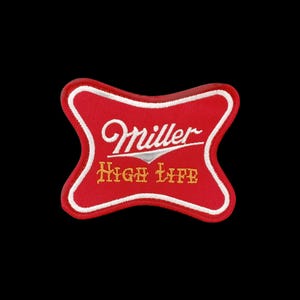 May include: A red embroidered patch shaped like a stylised hourglass with white borders. The text Miller is stitched in white script, and the words High Life are stitched in yellow block letters centred on the red fabric background.