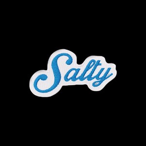 May include: An embroidered patch featuring the word Salty written in a cursive script font. The lettering is stitched in a bright blue thread, while the background of the patch is white with a thick white border outlining the entire shape.