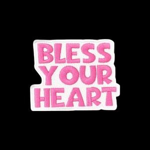 May include: A white embroidered patch with a die-cut shape featuring the text BLESS YOUR HEART in bold, pink, sans-serif capital letters. The embroidery has a visible textured thread pattern throughout the lettering and the white background.