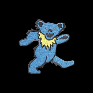 Pode incluir: Um emblema bordado apresentando um urso azul estilizado em uma pose dinâmica de caminhada. O urso tem um colarinho de pelo amarelo brilhante ao redor do pescoço, um nariz preto e uma boca larga e sorridente. O design é contornado por fio preto.