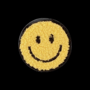 May include: A circular patch featuring a yellow smiley face design with a textured, fuzzy surface. The face has two black circular eyes and a wide black curved mouth, all set against a dark background with a defined black border edge.