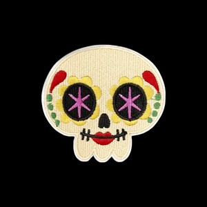 Puede incluir: Un parche bordado con forma de calavera y base de color crema. Los ojos son círculos negros con pétalos de flores amarillos y estrellas rosas en el centro. Presenta una pequeña nariz negra, labios rojos y puntos verdes en los laterales.