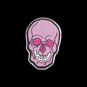 Puede incluir: Un parche bordado con forma de cráneo humano. El cráneo es de color rosa claro con las cuencas de los ojos y la cavidad nasal en rosa oscuro. Presenta una mandíbula blanca con dientes visibles y está delineado con un grueso borde bordado negro.