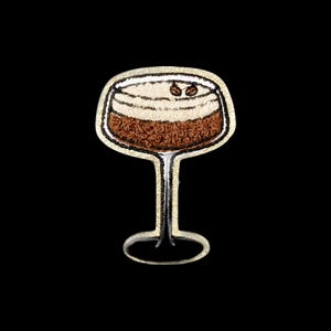 May include: An embroidered patch shaped like a cocktail glass filled with a brown liquid and topped with white foam. Two small dark brown coffee beans are placed on the foam. The entire design is outlined with a shimmering gold metallic thread border.