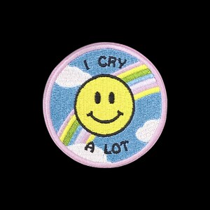 May include: A circular embroidered patch featuring a yellow smiley face in the centre against a light blue sky background with white clouds and a rainbow. The text I CRY A LOT is stitched in black letters above and below the smiley face.