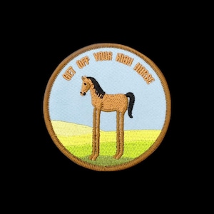 May include: A circular embroidered patch featuring a tan horse with long legs standing in a green field under a light blue sky. The text GET OFF YOUR HIGH HORSE is stitched in orange letters along the top curve of the patch, which has a brown border.