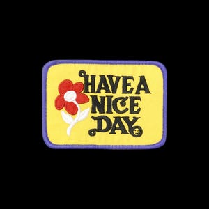 May include: A rectangular embroidered patch with a yellow background and a purple border. It features a red flower with a white stem and leaves on the left side. The black text reads HAVE A NICE DAY, with a small smiley face inside the letter Y.