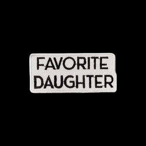 May include: A rectangular embroidered patch with a white background and a black border. The text "FAVORITE DAUGHTER" is stitched in bold, black, sans-serif capital letters across the centre of the patch, creating a clear and legible design.
