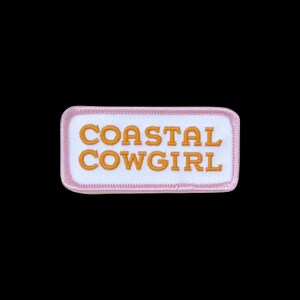 May include: A rectangular embroidered patch with a white centre and a light pink border. The text COASTAL COWGIRL is stitched in bold orange capital letters across the centre of the patch. The design features a clean, stitched edge finish.