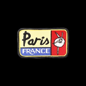 May include: A rectangular embroidered patch with a black border. The left side features the word Paris in black script above the word FRANCE in white block letters on a blue background. The right side shows a white ballerina on a red background.