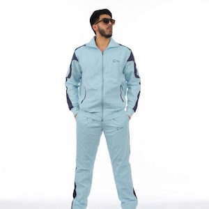 Könnte beinhalten: Hellblauer Trainingsanzug mit marineblauen Akzenten. Die Jacke hat einen Reißverschluss und die Hose einen geraden Beinverlauf. Der Anzug wird mit weißen Turnschuhen und Sonnenbrille getragen.