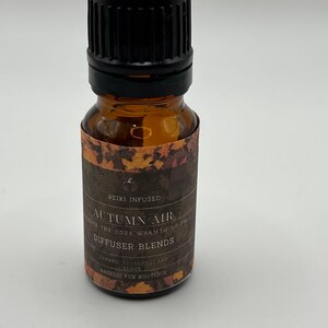 Peut inclure: Petite bouteille en verre ambré avec un bouchon noir. L'étiquette indique "Autumn Air Diffuser Blends" avec "Reiki Infused" en haut. L'étiquette présente un motif de feuilles d'automne et les mots "Orange, Patchouli and Clove".