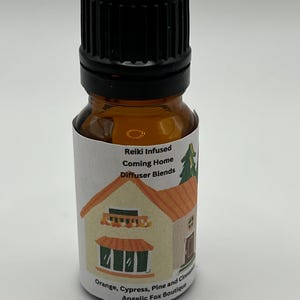 Può includere: Piccola bottiglia di vetro ambrato con tappo nero. L'etichetta presenta un'illustrazione di una casa e il testo "Reiki Infused Coming Home Diffuser Blends". Contiene arancia, cipresso, pino e cannella.