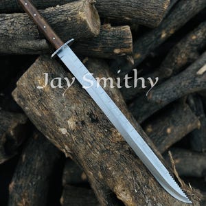 May include: A large, silver blade knife with a wooden handle and a silver guard. The knife is resting on a pile of wooden logs. The text "Jay Smithy" is printed on the blade.