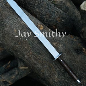 May include: A large sword with a silver blade, serrated on one edge, and a dark wooden handle. The text "Jay Smithy" is printed on the blade. The sword is displayed on a background of stacked logs.