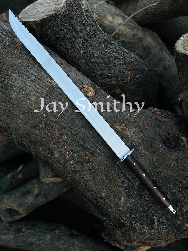 May include: A large sword with a silver blade, serrated on one edge, and a dark wooden handle. The text "Jay Smithy" is printed on the blade. The sword is displayed on a background of stacked logs.