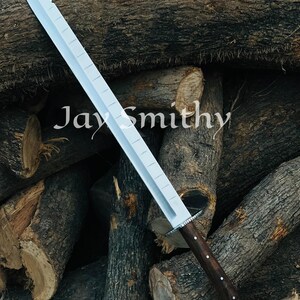 May include: A silver sword with a serrated edge and a dark wooden handle. The blade has a rectangular design with rectangular cutouts. The text "Jay Smithy" is visible on the blade. The sword is displayed against a backdrop of stacked logs.