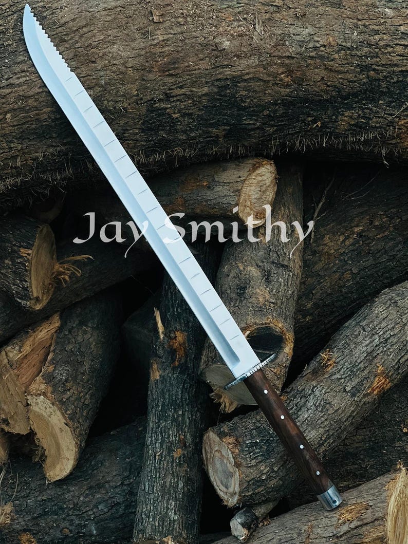 May include: A silver sword with a serrated edge and a dark wooden handle. The blade has a rectangular design with rectangular cutouts. The text "Jay Smithy" is visible on the blade. The sword is displayed against a backdrop of stacked logs.