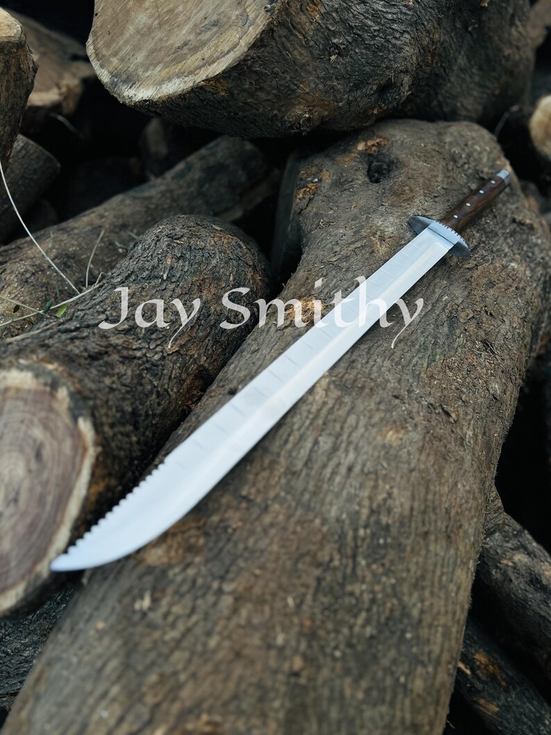May include: A large, silver sword with a serrated edge and a dark brown handle rests on a pile of wooden logs. The text "Jay Smith" is visible in the image.