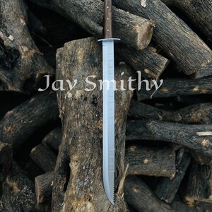 May include: A long, silver blade sword with a wooden handle. The handle is dark brown with small, light-coloured dots. The sword is resting on a tree stump, with the text "Jay Smithy" visible. The background is a pile of logs.