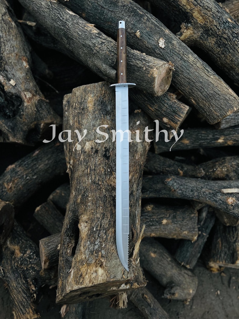 May include: A long, silver blade sword with a wooden handle. The handle is dark brown with small, light-coloured dots. The sword is resting on a tree stump, with the text "Jay Smithy" visible. The background is a pile of logs.