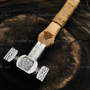 Può includere: Un martello artigianale con un manico in legno avvolto in spago. La testa del martello è color argento, con intricati disegni a nodo celtico. Il martello poggia su una superficie di legno scuro e strutturato.