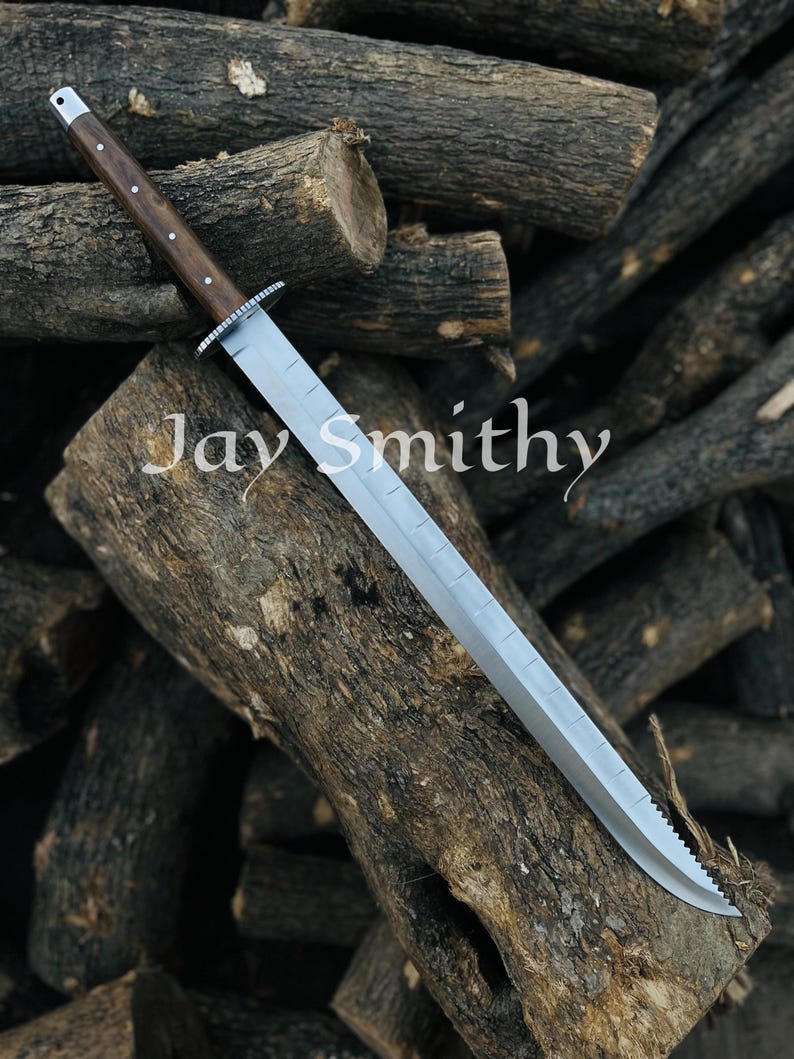 May include: A large, silver blade knife with a wooden handle and a silver guard. The knife is resting on a pile of wooden logs. The text "Jay Smithy" is printed on the blade.