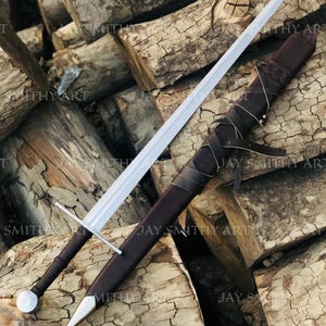 Könnte beinhalten: Ein langes, silbernes Schwert mit braunem Griff und passender Scheide. Das Schwert liegt auf einem Stapel Holzscheite. Die Scheide ist aus braunem Leder gefertigt und mit Lederriemen umwickelt.