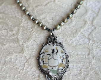 May include: A silver necklace with a large oval pendant featuring a black and white illustration of a woman in a garden setting. The necklace is made of faux pearls with a dark grey finish.