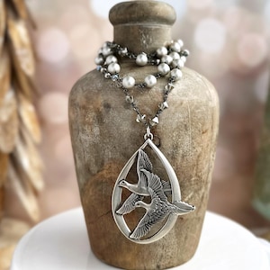 May include: A silver necklace with a teardrop-shaped pendant featuring two birds in flight. The necklace is made of silver beads and a chain with small silver accents.