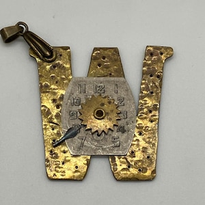 Puede incluir: Un colgante artesanal con forma de la letra "W". El colgante está hecho de latón con una textura martillada y presenta una esfera de reloj y un engranaje. Un pequeño dije en forma de pájaro está unido a la esfera del reloj.