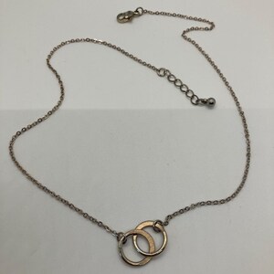 May include: A delicate silver-toned necklace featuring two interlocking gold-colored rings as a pendant. The necklace has a fine chain with an adjustable length and a lobster clasp closure. The pendant is a simple, elegant design.