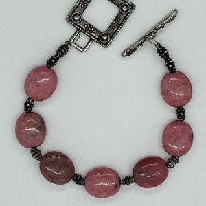 May include: A pink gemstone bracelet with a silver-tone clasp. The bracelet features oval-shaped pink stones strung together with small black beads. The clasp is a square shape with decorative detailing. A stylish piece of jewelry.