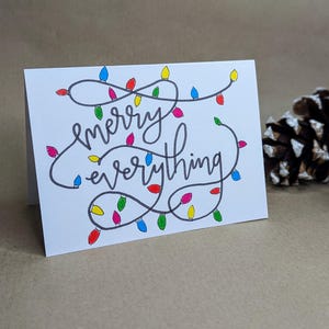 May include: A white card with the words "merry everything" written in black cursive. The words are surrounded by a string of colorful lights.
