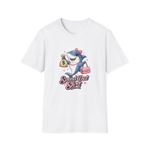 May include: A white short-sleeved t-shirt featuring a graphic of a cartoon shark wearing a pink bow and pearl necklace. The shark holds a tan money bag and a pink quilted handbag. Below the shark, the text reads "Spend Dat Sh*t" in pink.