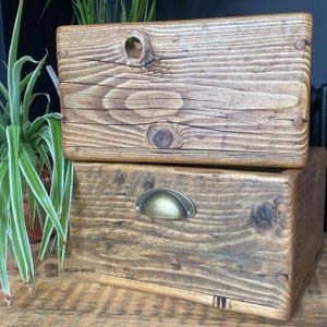 May include: Two stacked rectangular wooden storage boxes with a medium brown stained finish. The top box features a circular cutout handle, while the bottom box has a brass-coloured metal cup pull handle. Both boxes display visible wood grain.