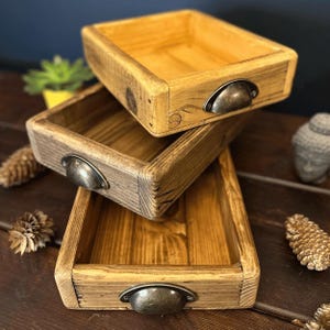 May include: Three wooden storage trays stacked. Each tray has a rectangular shape, rounded corners, and metal handles. The wood has a natural finish, with shades of brown and yellow. The trays are empty and ready for use.