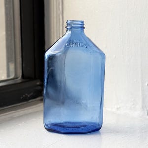 May include: A vintage blue glass bottle with the word "SQUIBB" embossed on the side. The bottle is empty and has a rounded rectangular shape.