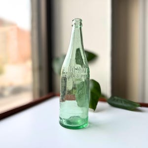 May include: A vintage, light green glass bottle with the embossed text "AUTO-MATIC AMERICA'S PHY". The bottle has a long neck and a rounded base. The bottle is displayed on a white surface near a window.