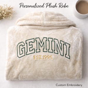 May include: A cream-colored plush robe with a hood. The robe features the word "GEMINI" embroidered in dark green, with "EST.1995" below it in gold. The text "Personalized Plush Robe" is above the robe, and "Custom Embroidery" is below.