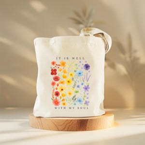 Puede incluir: Bolso tote de color crema con un dise&ntilde;o floral en colores del arco&iacute;ris. El texto "IT IS WELL WITH MY SOUL" est&aacute; impreso encima y debajo de las flores. El bolso tiene un asa de color natural y est&aacute; sobre un soporte de madera.