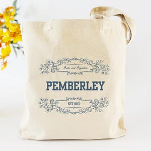 May include: Beige canvas tote bag with the text "PEMBERLEY" in navy blue, framed by decorative flourishes. The bag also features the text "Pride and Prejudice" and "EST 1813". The tote has a natural canvas handle.