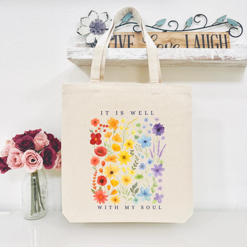 Puede incluir: Bolso tote de lona beige con el texto "IT IS WELL WITH MY SOUL" y un dise&ntilde;o floral colorido. Las flores est&aacute;n dispuestas en un degradado arco&iacute;ris, con flores rojas, naranjas, amarillas, verdes, azules y moradas. El bolso tiene dos asas.