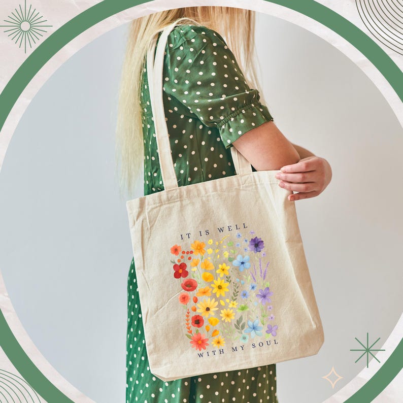 Puede incluir: Bolso tote de lona beige con un dise&ntilde;o floral colorido y el texto "IT IS WELL WITH MY SOUL". Las flores est&aacute;n dispuestas en un espectro de arco&iacute;ris. El bolso tiene correas color crema.