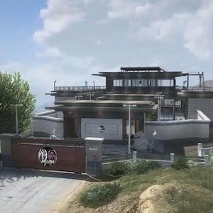 May include: A contemporary building with a flat roof and a gated entrance. The building is painted in shades of grey and white, with large windows and balconies. The gate features a circular design. The sky is blue with scattered clouds.