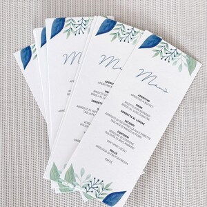Può includere: Un mazzo di biglietti di menu bianchi con un design ad acquerello di foglie blu e fogliame verde. La parola "Menu" è scritta in corsivo. I piatti sono elencati in italiano.