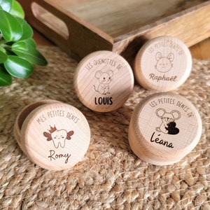 Peut inclure: Quatre petites boîtes rondes en bois avec des motifs gravés et du texte. Les boîtes présentent des illustrations de souris et du texte tel que "LOUIS", "Raphaël", "Léana" et "Romy". Les boîtes sont sur une surface tissée.